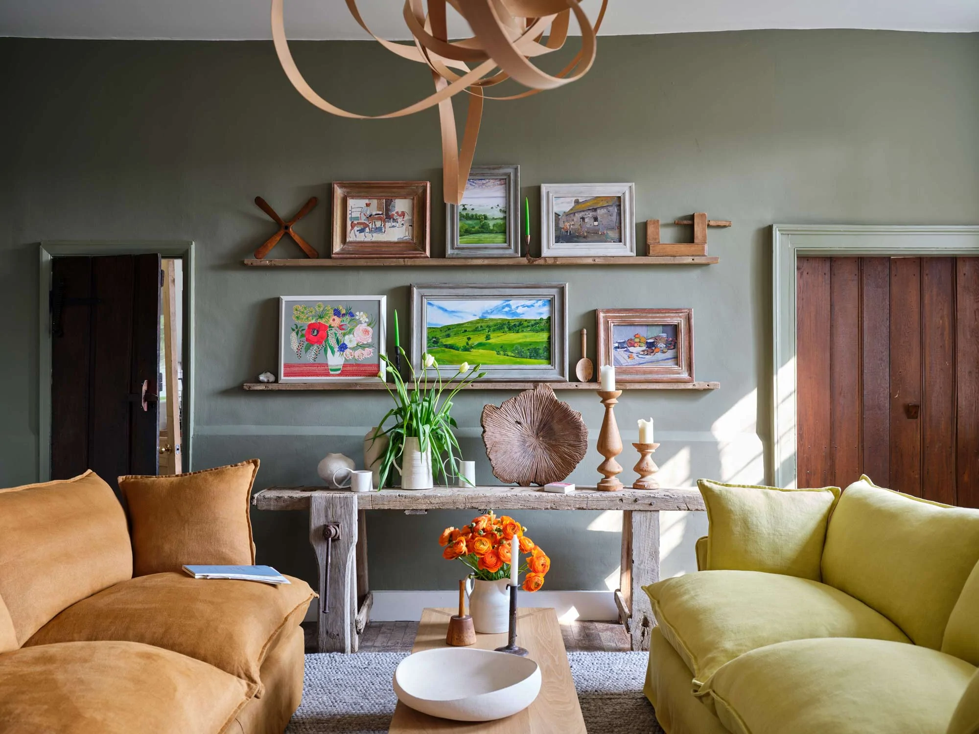 A soft, warm moss green room with a citrine yellow and brown bronzite sofa facing each other with a birch coffee table in between. The walls are covered in paintings and decorations in 3 horizontal rows. Against the wall is a rustic wooden side table with candles, a vase of white tulips and a wooden sculptural bowl.